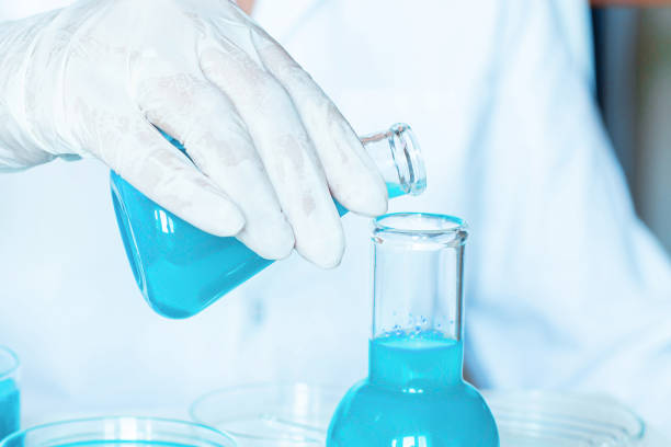 istock Lab Locale: Flask Grasped by Assistant, Pouring Blue Liquid into Bottle. Ivory Attire. Glass Equipment. 1636608148