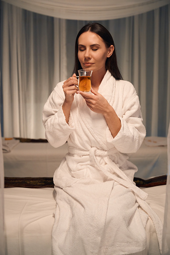 Spa customer dressed in bathrobe sitting on couch and inhaling aroma of drink in her hands