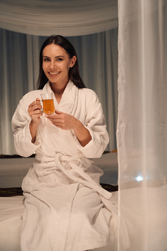 Smiling young woman in terry bathrobe sitting on massage couch with cup of tea in hands