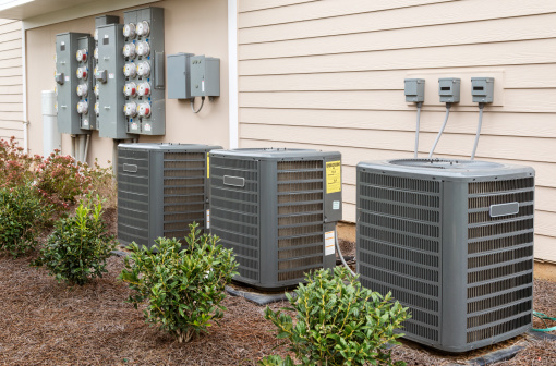 Three air conditioning units with a panel of electricity meters behind, all at the side of a recently-constructed apartment building.  The yellow energy usage stickers are not from the manufacturer, but the U. S. government.