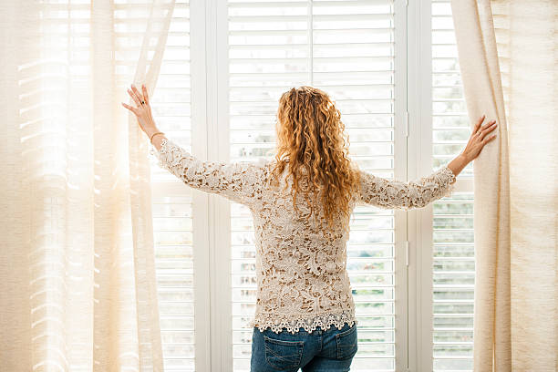 Woman looking out big bright window with curtains and blinds