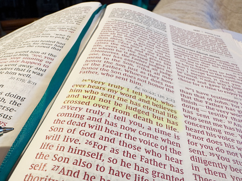 Bible open to John 5:24. Text is marked in yellow highlighter. Teal ribbon bookmark runs down the center of the bible. Jesus’ words are in red letters.