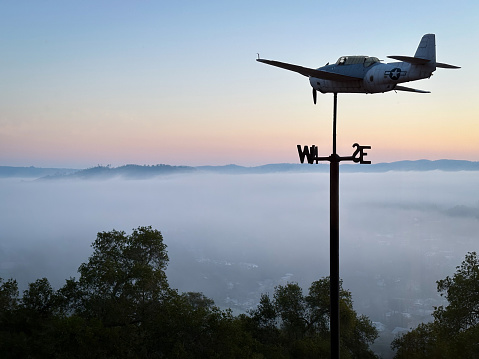 Looking at fog filled valley with trees and airplane type weather vane silhouetted against glowing sunrise in the early morning