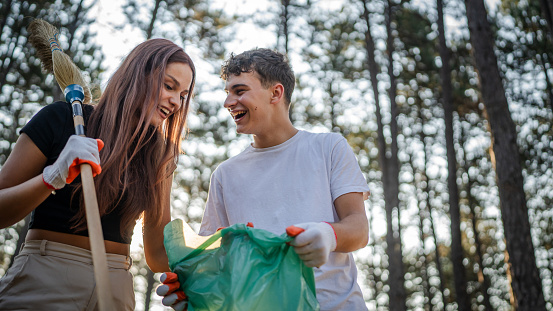 couple or teenage friends gen Z male and female caucasian man woman picking up waste garbage plastic bottles and paper from forest cleaning up nature in sunny day environmental care ecology concept