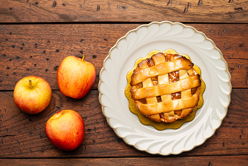 High angle view of three apple fruit and delicious apple pie on a white plate with a yellow carton base over a rustic wood table in the kitchen at home ready to be eat. Image made in studio