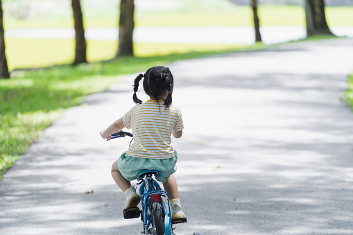 Back side view of asian baby girl child learning to ride bicycle in the park garden. Education concept for kid practiice cycling at park, baby sport concept.