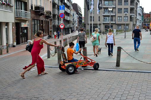 Blankenberge, West-Flanders, Belgium - August 24, 2023: North-African mother red dress helping sun sitting in a rented go-cart to get from street on footpath square