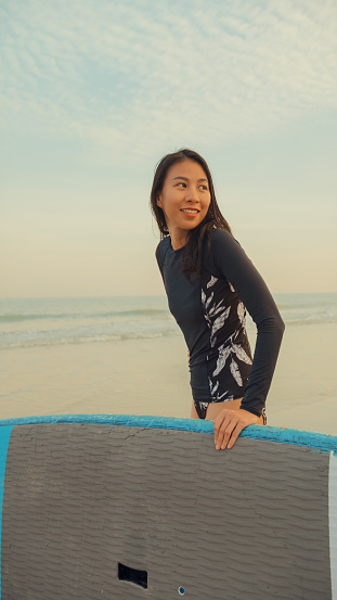 Young Asian woman surfer with surfing board on tropical beach. Healthy active lifestyle and summer vacation concept. Vertical Screen.