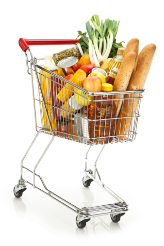 Three quarters view of a metal shopping cart filled with a large variety of colorful groceries that includes some fresh vegetables, fruits, canned food, fruit juice, cooking oil and three loafs of bread. The cart is standing on reflective white background producing a soft reflection under it. The cart has a red plastic handle. DSRL studio shot with Canon EOS 5D Mk II