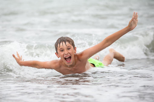 A cheerful happy boy plays with sea waves, spreads his arms and screams with delight.