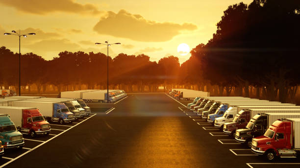 A tranquil and atmospheric scene of a parking lot at dusk, just after a rain shower. The wet asphalt reflects the warm, vibrant colors of the setting sun, which paints the sky in a stunning gradient of deep purples, pinks, and soft oranges. A row of tall streetlights casts a bright glow on the pavement, contrasting with the fading natural light. In the foreground, shopping cart corrals and a few parked cars add a sense of everyday life, while the silhouettes of trees and buildings anchor the horizon. A faint crescent moon is visible in the deepening sky, completing this peaceful urban twilight view. This image is perfect for themes of end-of-day, urban life, mood, weather, and quiet moments in a commercial environment.