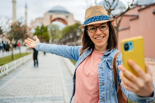 Cheerful young woman taking selfies in front of Hagia Sophia Grand Mosque (Ayasofya Camii) in Istanbul, Turkey.