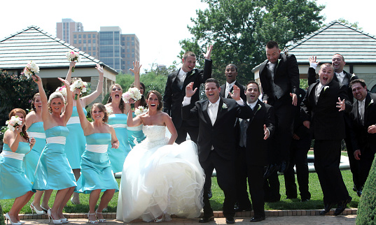 Wedding portrait of a bride, groom and wedding party smiling and waving with the view of city buildings in the background.  The woman in a white wedding gown and man in a black tuxedo are standing in the center of the image with eight men in tuxedos lined up in a row to the right of the image.  Seven women in teal strapless dresses with a white sash are lined up to the right of the bride.  The wedding party members have their hands in the air and the young women are holding bouquets of flowers.  City buildings, grass and a concrete sidewalk are in the background.