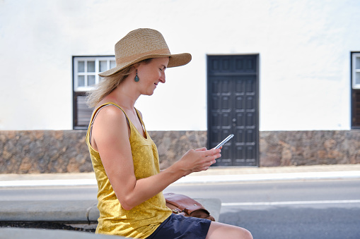 Side view of satisfied woman in straw hat browsing cellphone while chilling on bench near building in sunlight