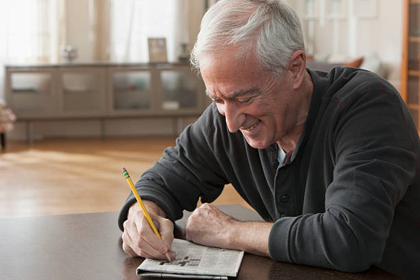 A senior man working on a crossword puzzle in his home Smiling senior caucasian man working on crossword puzzle, horizontal view crossword stock pictures, royalty-free photos & images