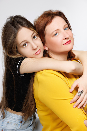 Portrait of naturally blond teenage girl embracing her middle aged mother from behind, they are looking at camera, Mother's Day celebration, studio shot