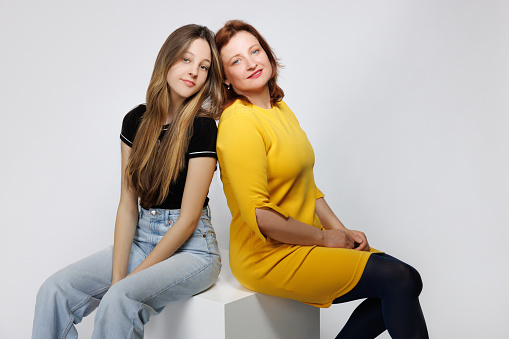Middle aged woman with dyed red hair sitting back to back with her naturally blond teenage daughter, they are looking at camera, Mother's Day celebration, studio shot
