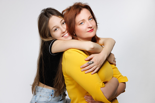 Portrait of happy naturally blond teenage girl embracing her middle aged mother from behind, they are looking at camera, Mother's Day celebration, studio shot