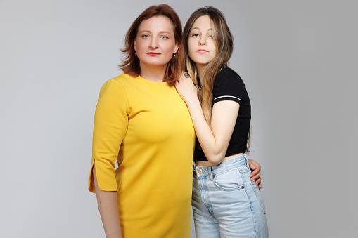 Portrait of middle aged woman with dyed red hair standing close to her naturally blond teenage daughter embracing her, they are looking at camera, Mother's Day celebration, studio shot