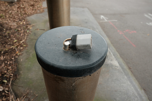 Silver padlock attached to a metal bollard by the side of a road.