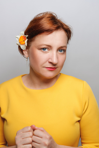 Portrait of mature woman with dyed red hair and blue eyes, holding hands on chest, daffodil flower behind her ear and looking at camera, studio shot