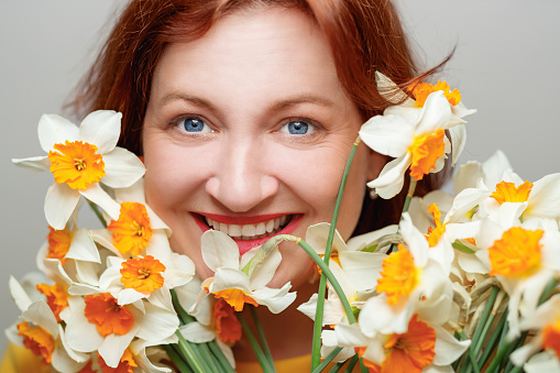 Portrait of mature woman with dyed red hair and blue eyes, smiling happily at camera from behind a bunch of daffodils, studio shot