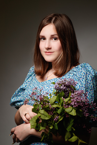 Portrait of beautiful teenage girl with brown hair in spring dress looking at camera seriously and holding bunch of lilac, studio shot