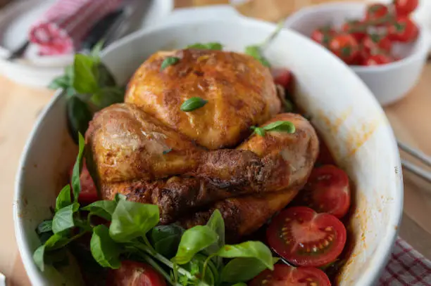 Oven baked crispy whole chicken with cherry tomatoes and basil. Served hot and ready to eat in a baking pan on a dinner table.