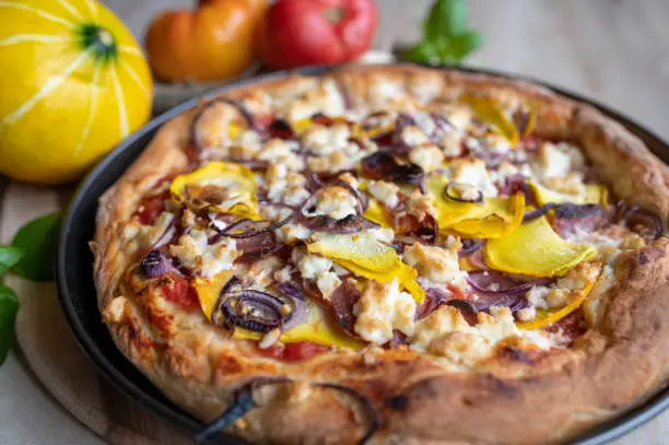 Homemade fresh baked pizza with goat feta cheese, spanish salami, yellow pumpkin, red onions and tomatoes, Served hot and ready to eat in baking pan. Closeup