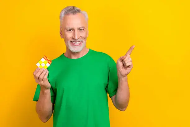 Photo of Photo of good mood positive man wear green t-shirt holding cube game pointing empty space isolated yellow color background