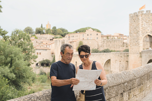 Older tourist couple sightseeing on city street with map - Happy husband and wife enjoying summer vacation together - Tourist lifestyle concept with older woman and man traveling around Besalu, Catalonia.