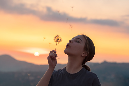 Portrait of a nice teen girl over a beautiful sunset sky in the mountains. Having fun and blowing on the dandelion flower. Making a wish. Happy, joyful, active teenage age.