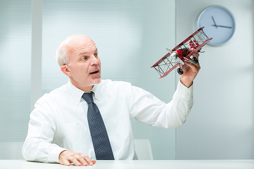 Older man, feeling youthful, plays with a red tin toy airplane on his work desk. Perhaps a visionary leader, he seeks inspiration through such leisure