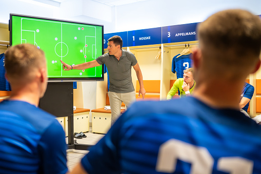 Football coach pointing while explaining game strategy on device screen to football players in dressing room.