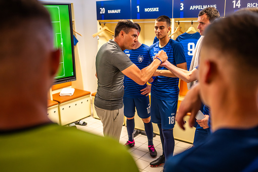 Male soccer players team listening to coach explaining game strategy in locker room before match, selective focus