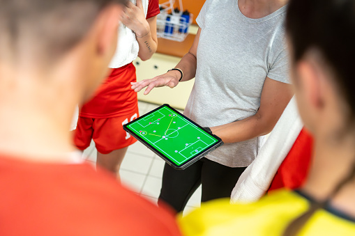 Female football coach explaining game strategy on digital tablet to football players in dressing room.