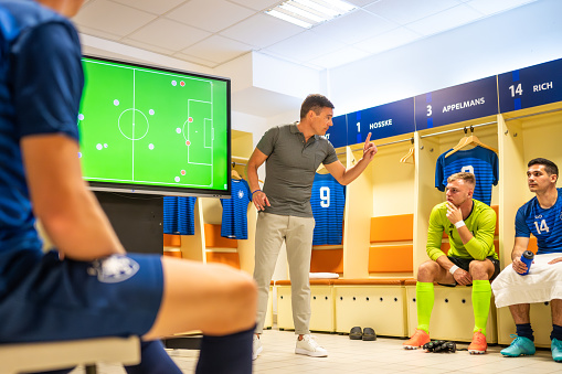 Football coach looking away while explaining game strategy on device screen to football players in dressing room.