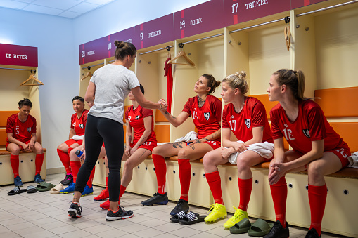Female coach holding hands while talking to female football player sitting in dressing room.
