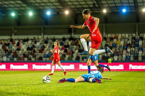 Female football player jumping mid-air over opponent teammate lying on grass during match on football pitch.