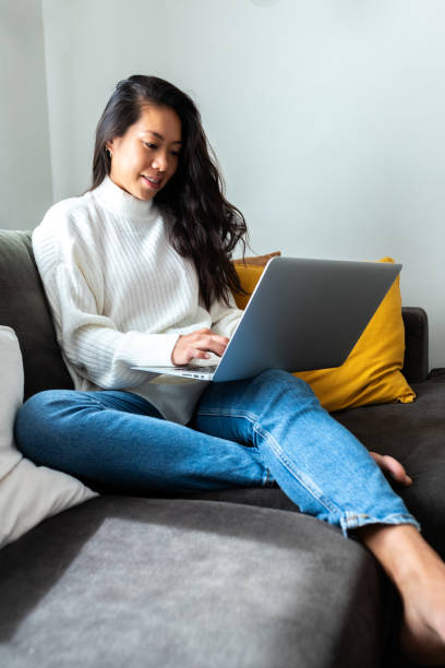istock Young Asian woman using laptop at home sitting on the couch, relaxing. Vertical image. 1637743291