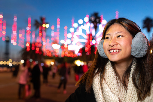Happy young Asian woman wearing ear muffs and scarf on a cold winter night looking away. Copy space. Lifestyle concept.
