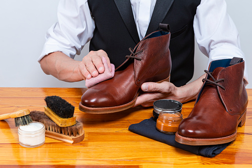 Professional Male Shoes Cleaner with Cleaning Brushes For Tan Derby Leather Boots While Working in Workshop With Rub Cloth. Horizontal Image