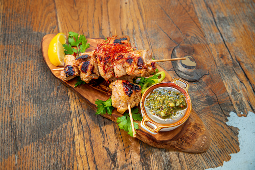 Juicy chops of chicken meat on skewer, grilled and served on wooden board with herbs in oil and lemon slice, garnished with chili threads and parsley, wooden table in fine dining