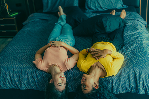 Lesbian lovers engage in playful interactions on bed. Their eyes meet with an affectionate gaze, radiating love and tenderness. Laughing, sharing intimate moments filled with warmth and playfulness.