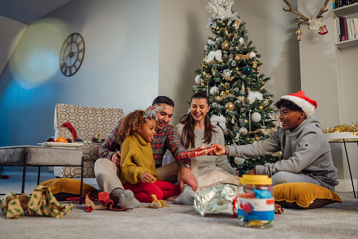 A media wide angle view of a young family exchanging gifts in their living room. They are dressed in festive attire and look cheerful. Behind them is their decorated xmas tree. The father is sitting with his daughter as they pass gifts to each other.