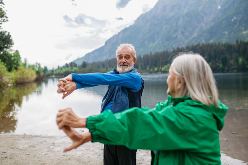 Senior couple doing outdoor yoga, tai chi, pilates by the lake in the autumn. Elderly husband and wife spending active vacation in the mountains, enjoying combination of physical activity and relaxation. Pensioners stretching after outdoor workout