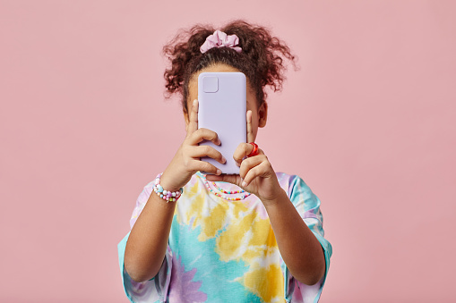 African American girl holding smartphone in front of her face and looking at screen while taking selfie or communicating in video chat
