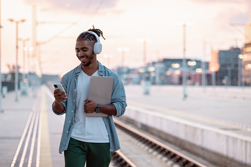 Smiling male student holding laptop and smart phone. He is waiting for a train on a train station at dusk