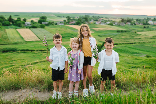 The happy children with flowers in a field and mountains on a sunny summer day at sunset. Cute kids laugh and walk on green grass in the spring.