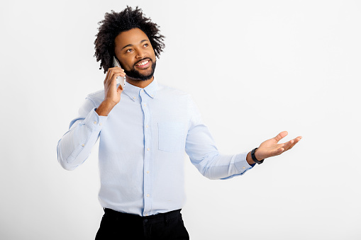 Always in touch. African-American curly man in white formal shirt talking on the smartphone and gesturing, having conversations with customers or colleagues, studio shot isolated on white